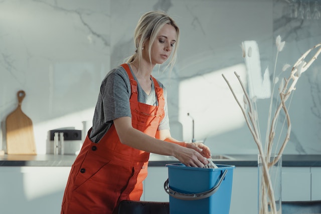 Person in orange coveralls soaking a rag in a bucket in a kitchen